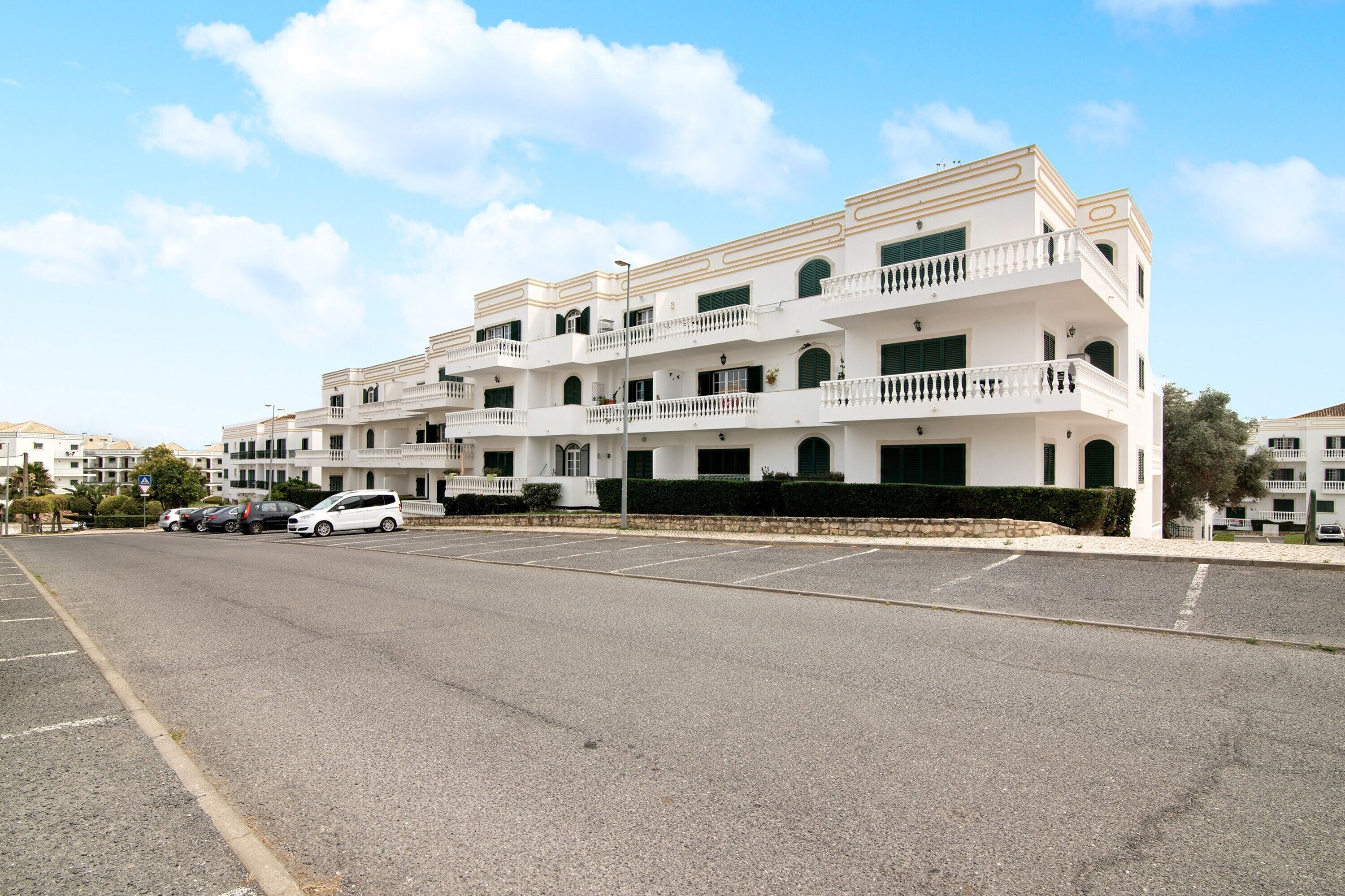 Un edificio blanco de varios pisos con balcones, un estacionamiento y un cielo despejado.