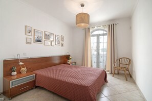 Une chambre à coucher avec une tête de lit en bois, un lit recouvert d’une courtepointe rose à motifs, une lampe de chevet, un fauteuil en osier et une grande fenêtre avec des rideaux.