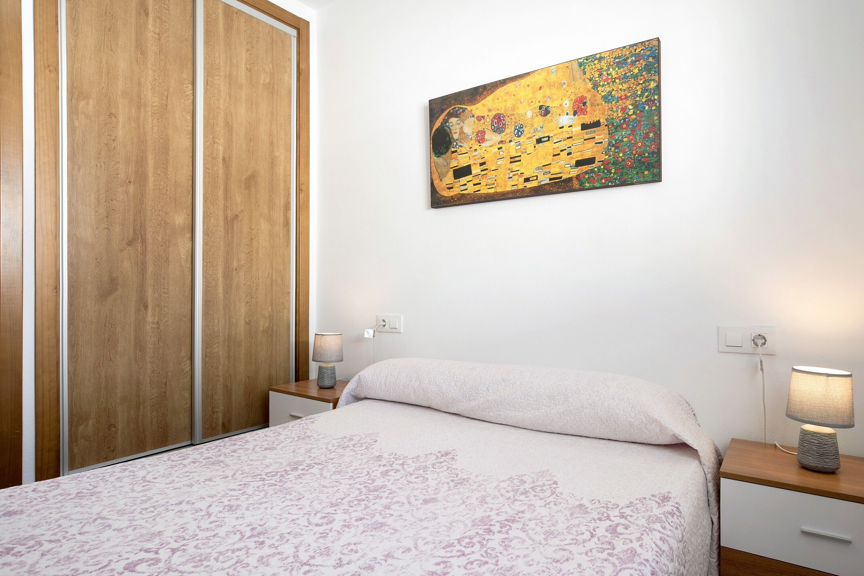 Un dormitorio con cama, mesitas de noche, un cuadro colgado en la pared y un armario de madera.