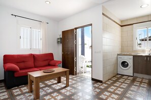 Un espace de vie compact comprenant un canapé rouge, une table en bois, un lave-linge et un sol carrelé.