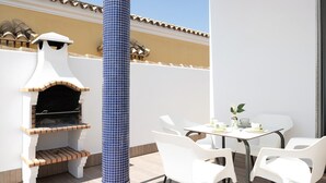 Un patio al aire libre con un horno de ladrillo, sillas blancas y una mesa con un florero.