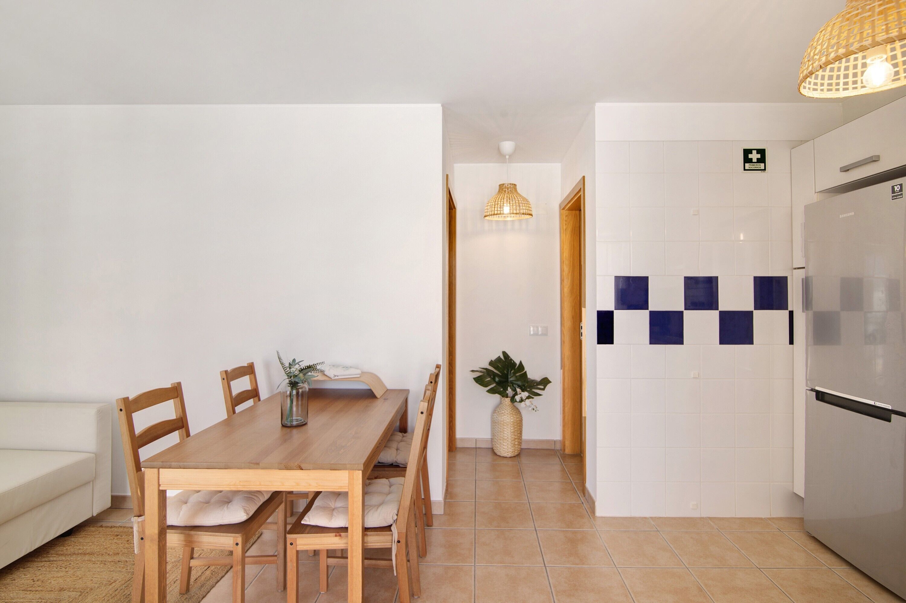 A dining area with a wooden table and chairs, a sofa, a hanging light fixture, and a refrigerator.