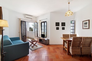 A living room with a sofa, dining table, chairs, a TV, and a hanging light fixture. - Vacation Home 'Quinta Velha Village 134" with Balcony, Shared Pool & A/C (Cabanas de Tavira)