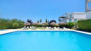 En swimmingpool med liggestole og parasoller, omgivet af grønt og en moderne bygning.