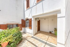 Un bâtiment blanc avec une cour dallée, des volets en bois et des plantes en pot.