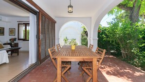 A wooden dining table with chairs, a potted plant, and a hanging light fixture in a covered outdoor area with a view of greenery.