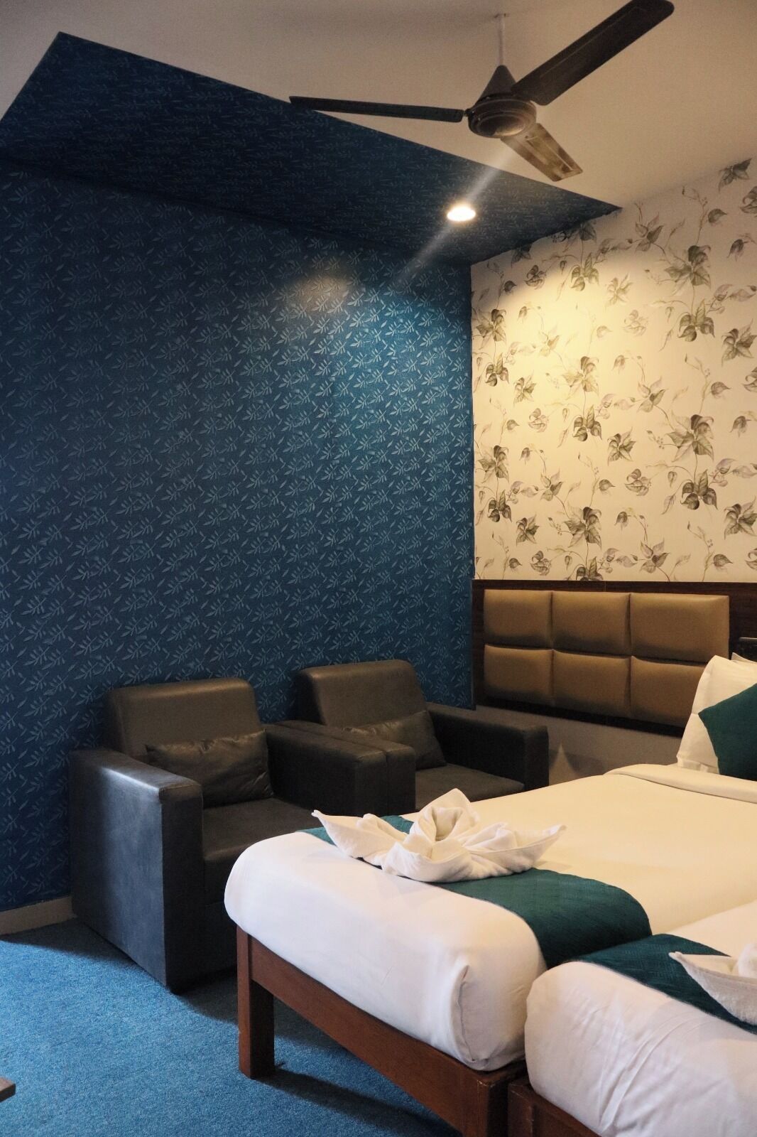 Una habitación de hotel moderna con una pared azul texturizada, un panel de papel tapiz floral, una cama con ropa blanca y dos sillones de cuero.