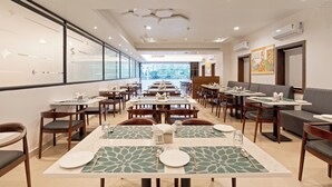 A modern dining area with multiple tables and chairs, a large window offering a view, and a ceiling with recessed lighting. - The Elite Hotel  (Hyderabad)