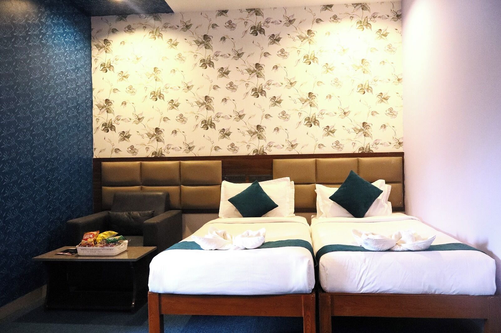Habitación de hotel con dos camas, un sofá, una mesita con aperitivos y papel tapiz con motivos florales.