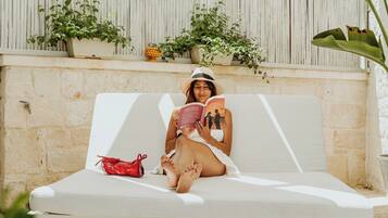 Una persona sentada en un banco blanco al aire libre, con un libro, rodeada de plantas en macetas y una pared blanca.