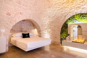 A bedroom with a stone wall, a bed with white bedding, and a small fireplace. - La Mandorla Luxury Trulli (Alberobello)