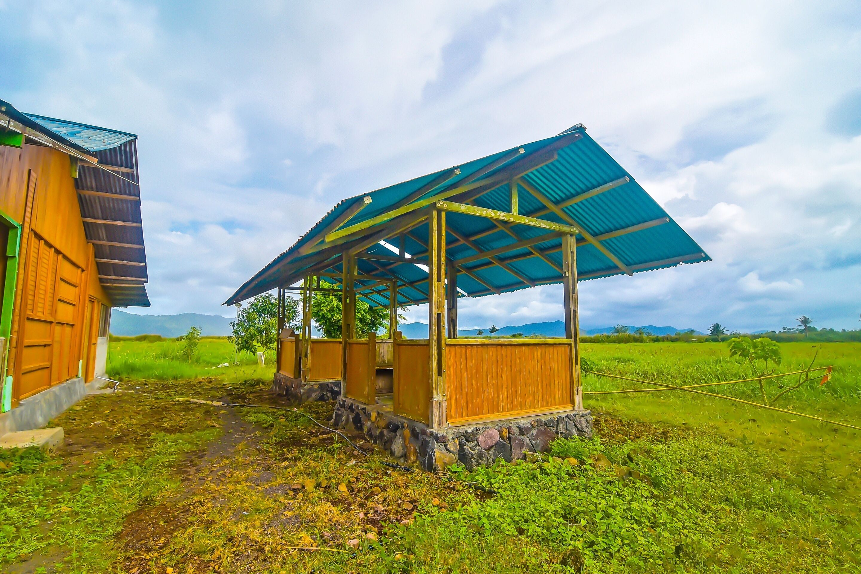 A rustic wooden structure with a corrugated metal roof, surrounded by a lush green field and distant mountains.