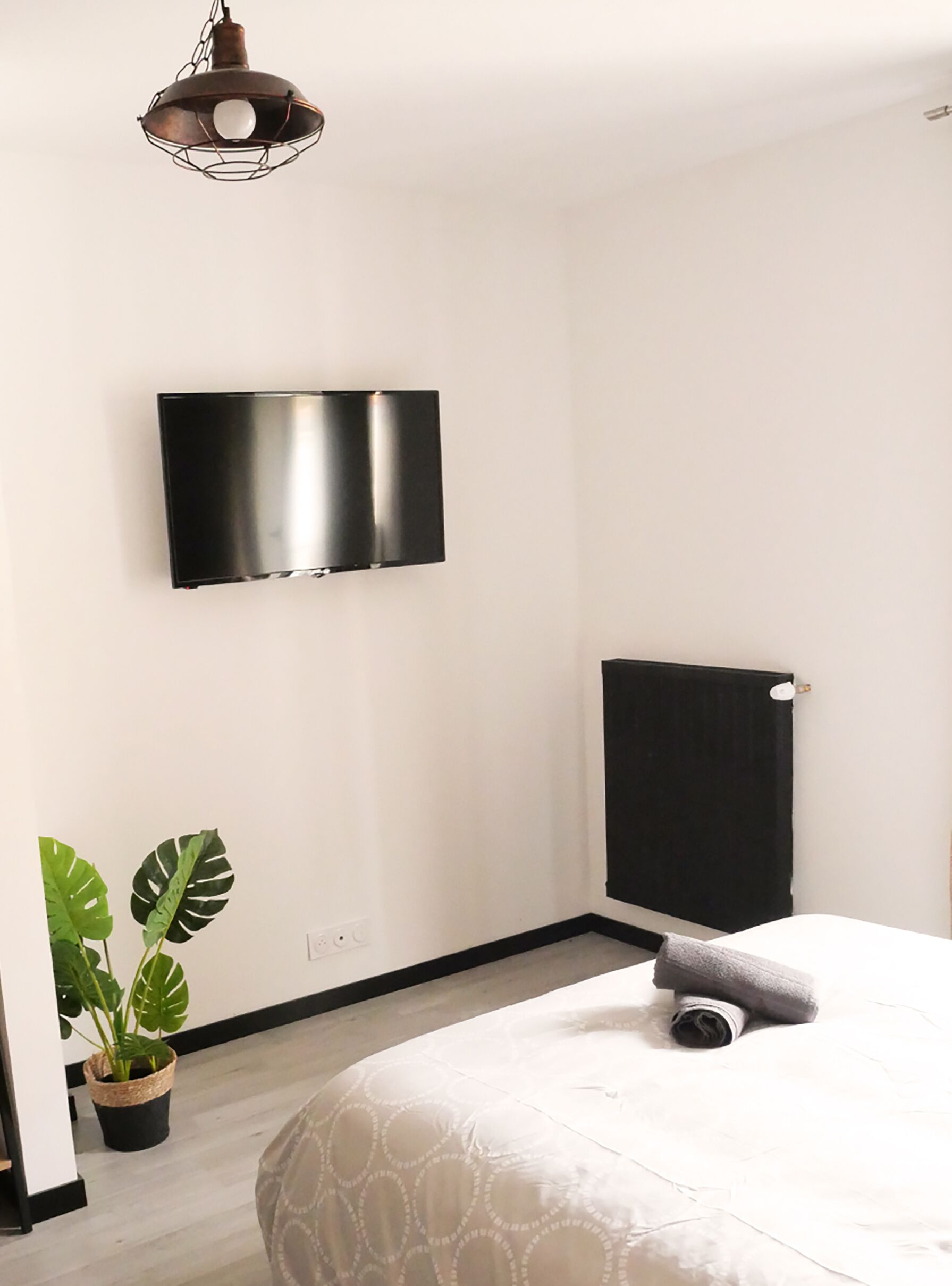 A bedroom with a bed, a potted plant, a flat-screen TV mounted on the wall, and a radiator.