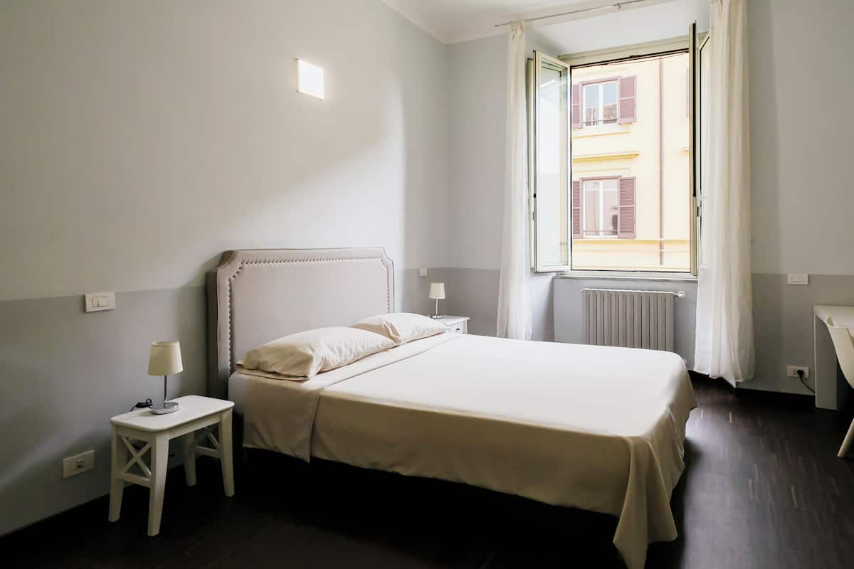 Ein Schlafzimmer mit Bett, Nachttisch mit Lampe, einem Fenster mit Vorhängen und einer Heizung.