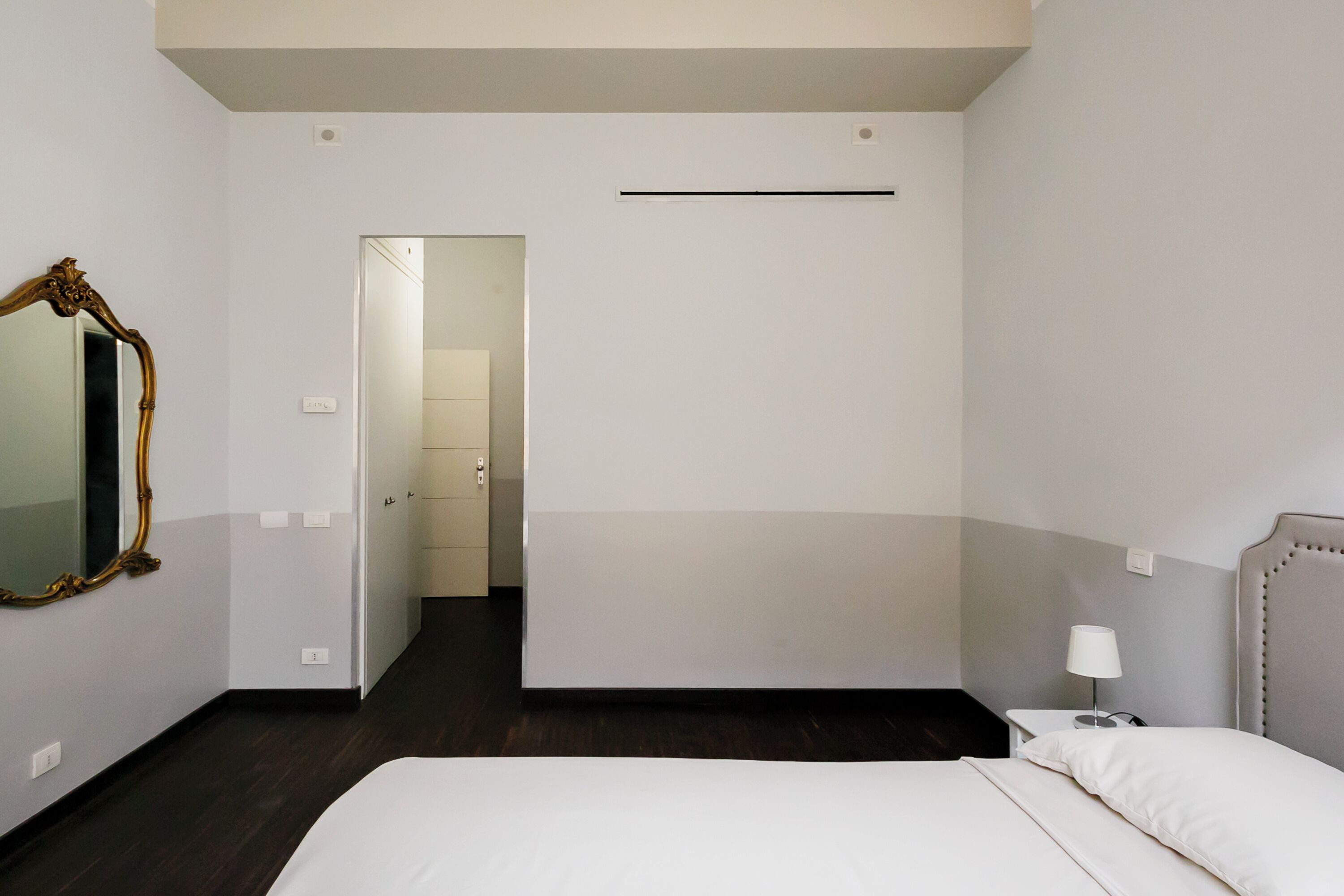 Una habitación de hotel con cama, mesita de noche, lámpara, espejo y una puerta que da a otra habitación.