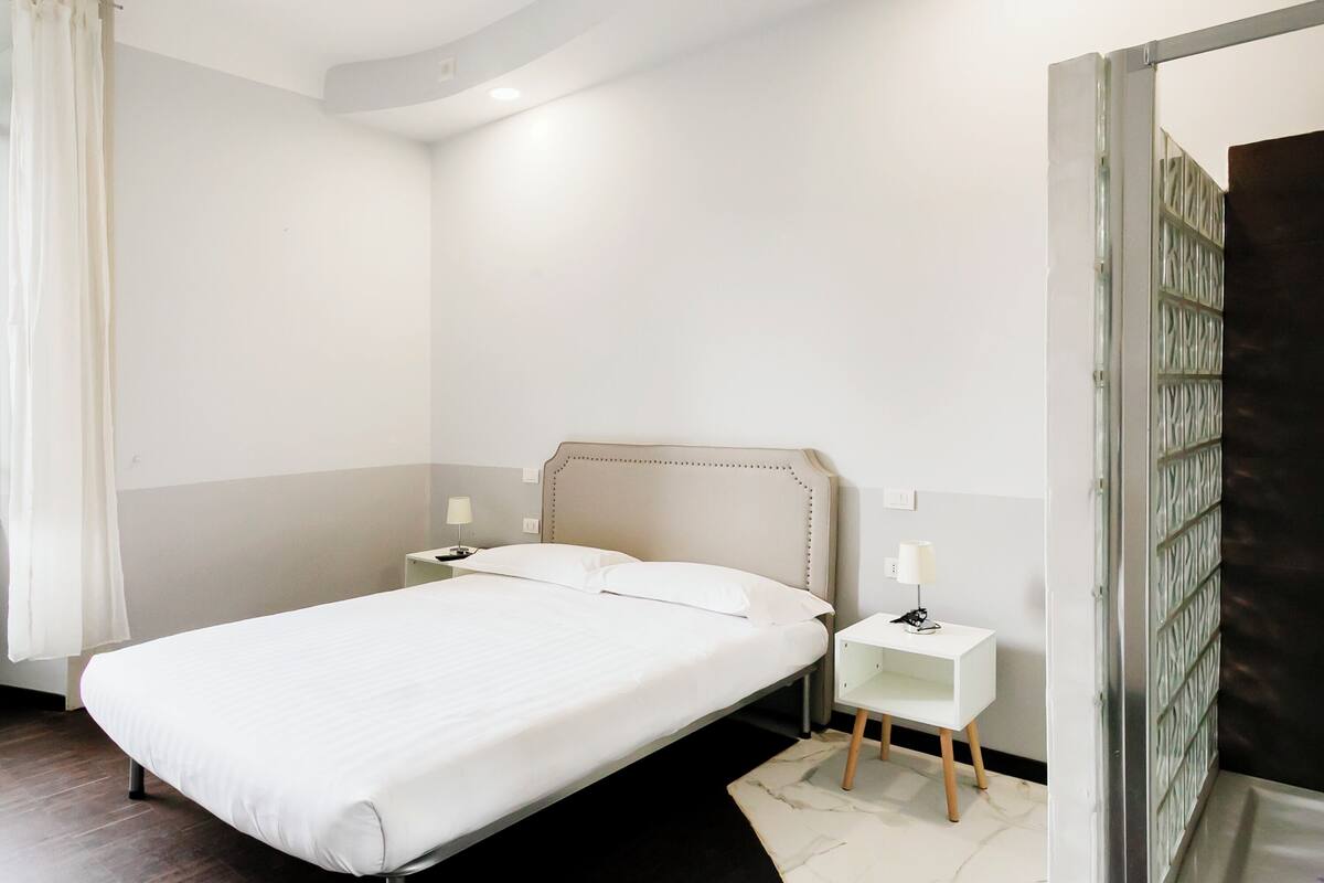 Ein modernes Hotelzimmer mit einem großen Bett, Nachttischen und Blick auf einen abgedunkelten Raum durch eine Glastür.