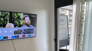 Una sala de estar con una televisión de pantalla plana montada en la pared, un sofá con cojines y un ventanal corredero que da a un balcón exterior.