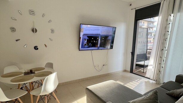 Una sala moderna con un televisor de pantalla plana montado en la pared, un comedor con una mesa y sillas blancas, y un sofá gris.