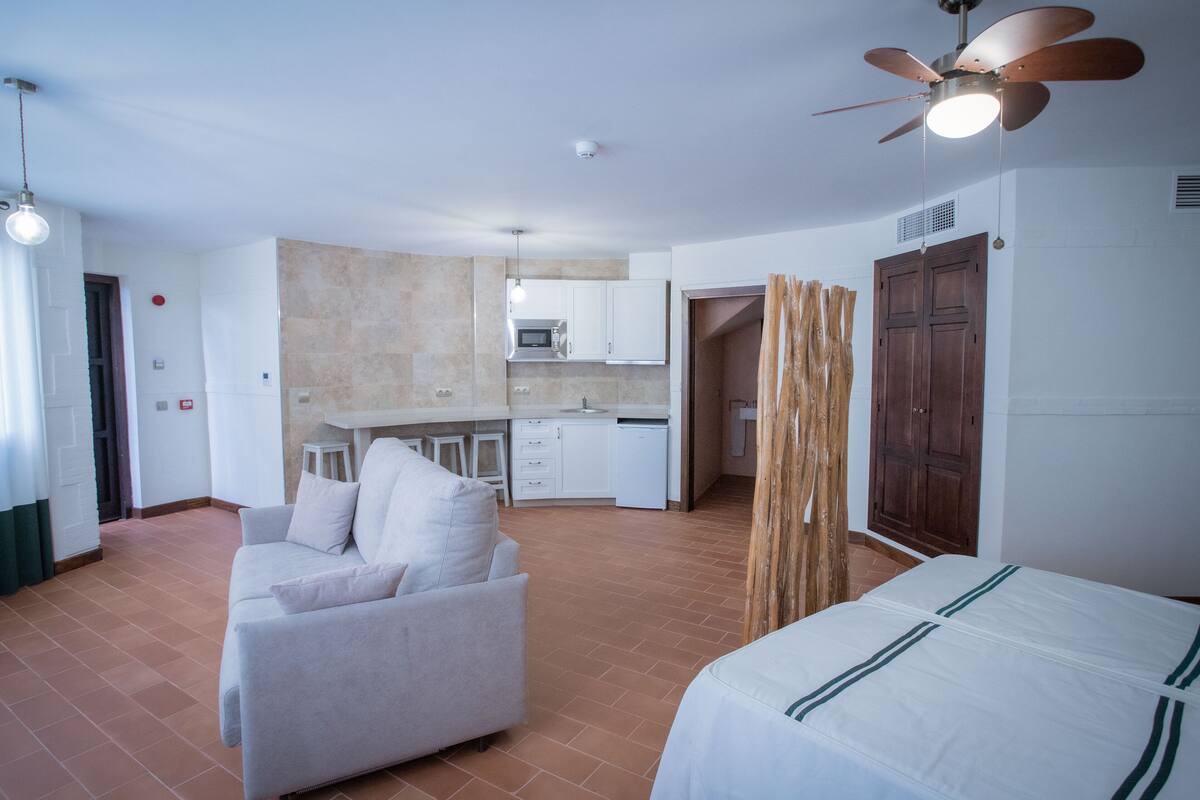 Una habitación de hotel moderna con sofá, cama, ventilador de techo y una kitchenette al fondo.