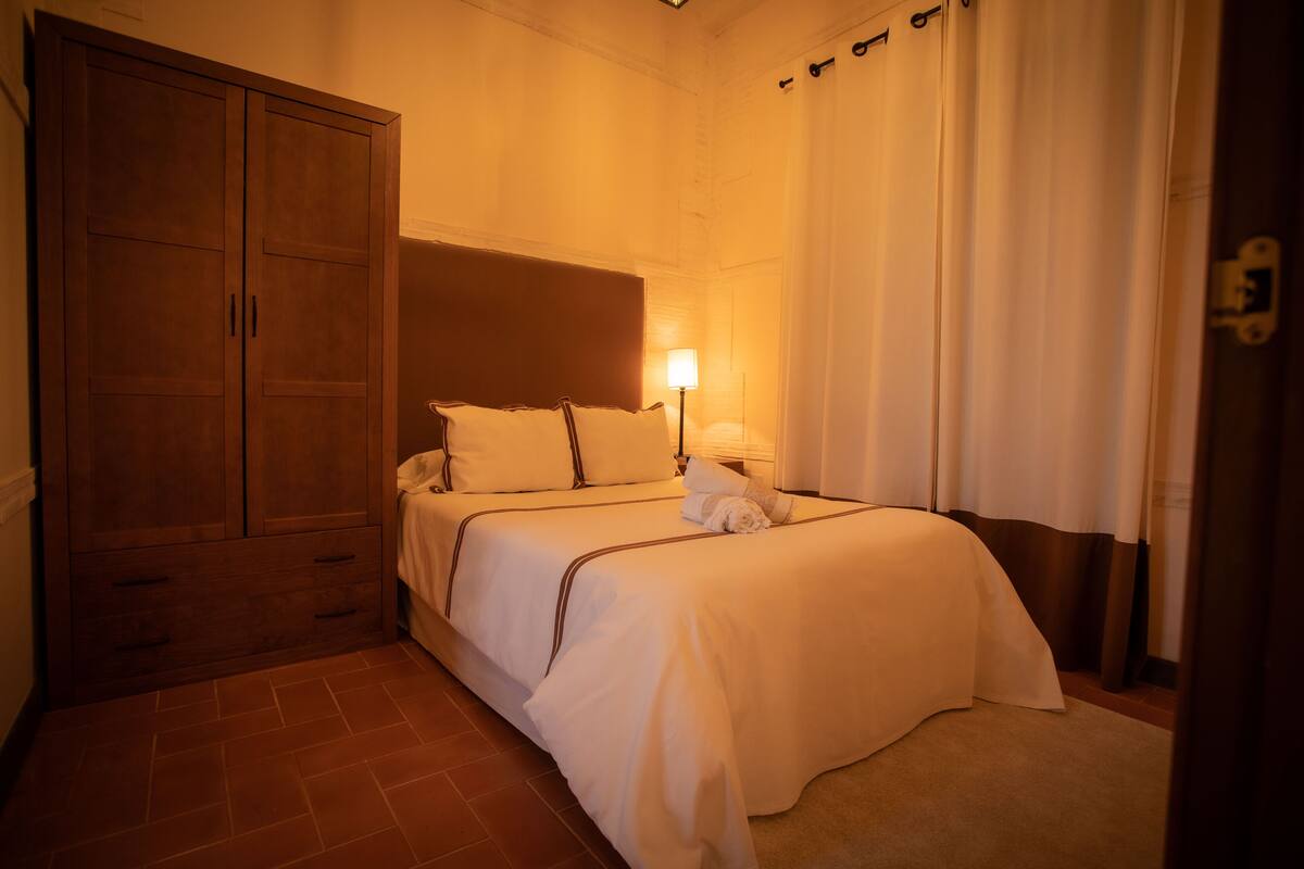 Habitación de hotel con un armario grande de madera, una cama con ropa blanca, una lámpara de noche y una ventana con cortinas.
