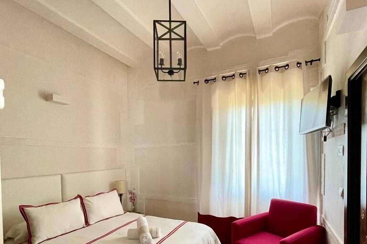 Habitación de hotel con una cama grande, un sillón rojo, una lámpara en el techo y una ventana con cortinas.