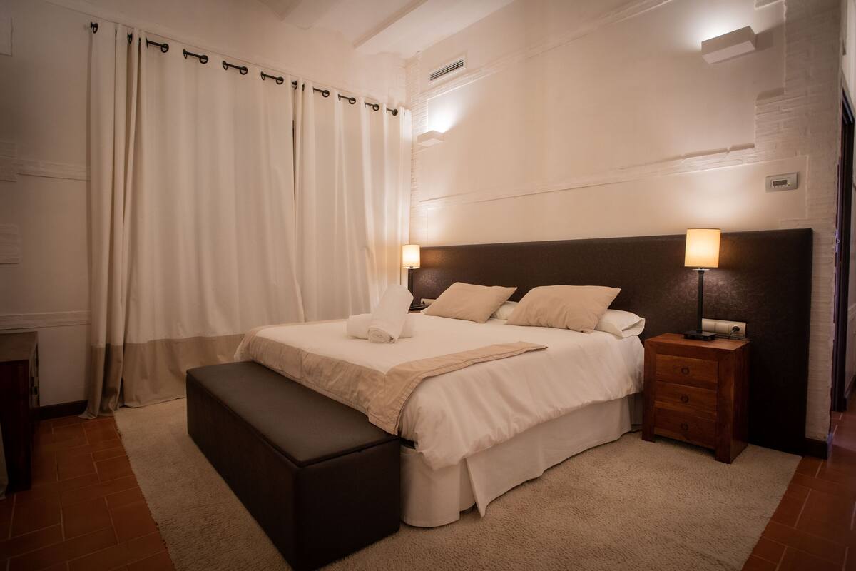 Un dormitorio con una cama grande, una mesita de noche con lámpara, un banco y una cortina.