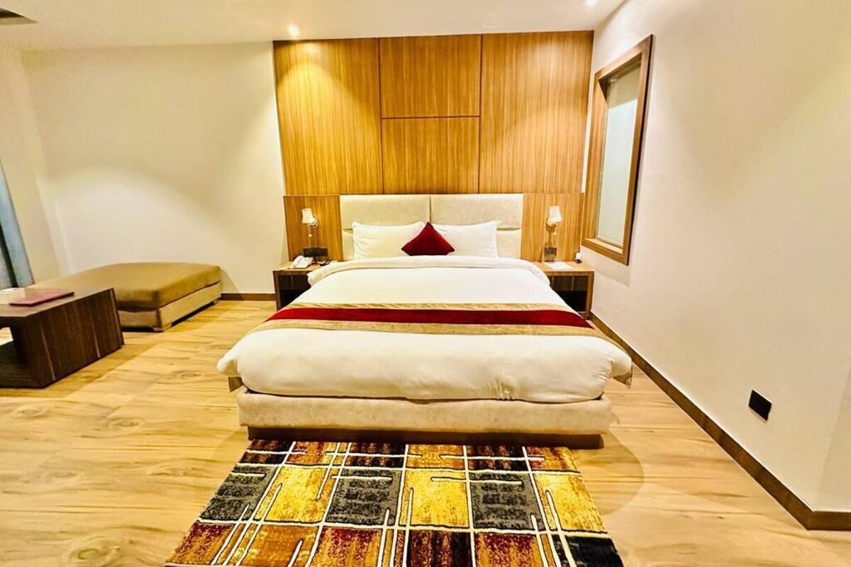 A modern hotel room with a large bed, wooden paneling on the walls, a bedside table with a lamp, and a view of the city.