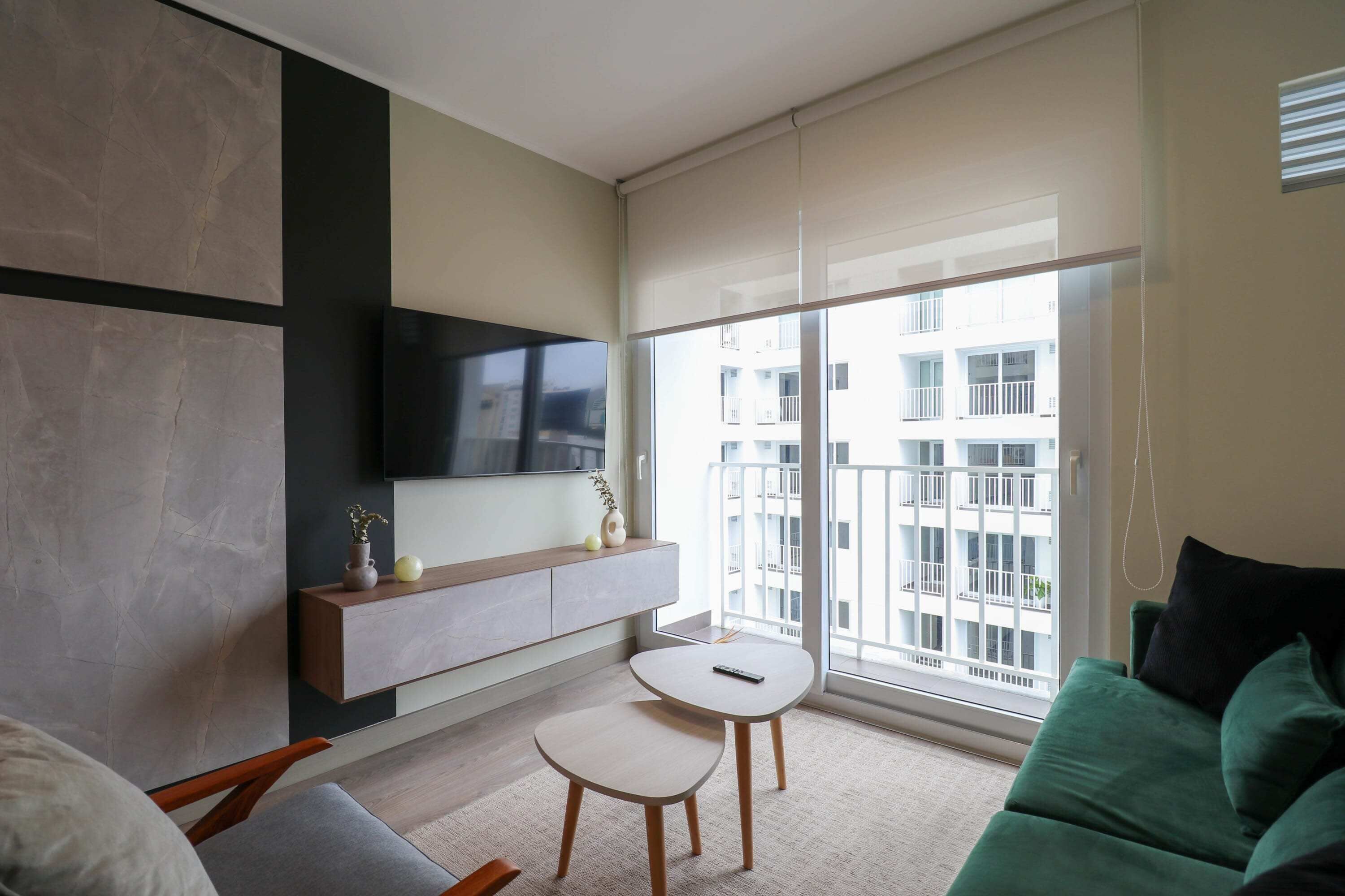 A modern living room with a sofa, a small table, a mounted TV, and a large window with a view of a building.