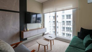 A modern living room with a sofa, a small table, a mounted TV, and a large window with a view of a building. - w* | Spectacular 2BR in the Heart of Miraflores (Miraflores)