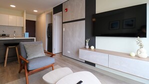 A modern living room with a grey sofa, a white ottoman, and a wooden coffee table. A flat-screen TV is mounted on the wall, and there is a small white vase with flowers on a shelf. - w* | Spectacular 2BR in the Heart of Miraflores (Miraflores)