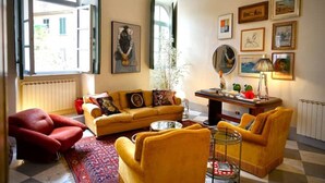 Ein Wohnzimmer mit gelbem Sofa, rotem Sessel und Couchtisch. An der Wand hängen Bilder in Rahmen und auf dem Tisch steht eine Pflanze.