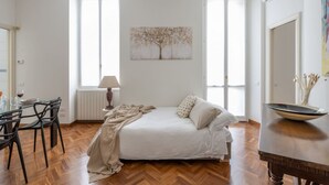 Een slaapkamer met een bed, een eettafel, stoelen, een radiator en een schilderij van een boom.