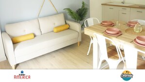 Ett modernt vardagsrum med en beige soffa, gula kuddar, ett matbord dukat med rosa tallrikar och vita stolar. En flätad korg hänger på väggen och en krukväxt syns.