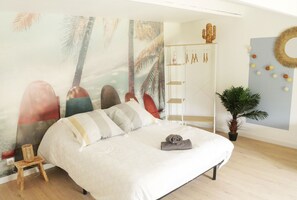 Ein Schlafzimmer mit einem Bett, einem kleinen Hocker, einem an der Wand befestigten Regal mit Pflegeprodukten, einer Topfpflanze und einem Wandbild mit Palmen.