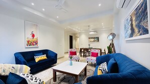 A modern living room with two blue sofas, a coffee table, and a dining area with a table and chairs. - BluO 3BHK CityWalk Mall, MAX Saket -Lift, Balcony (New Delhi)