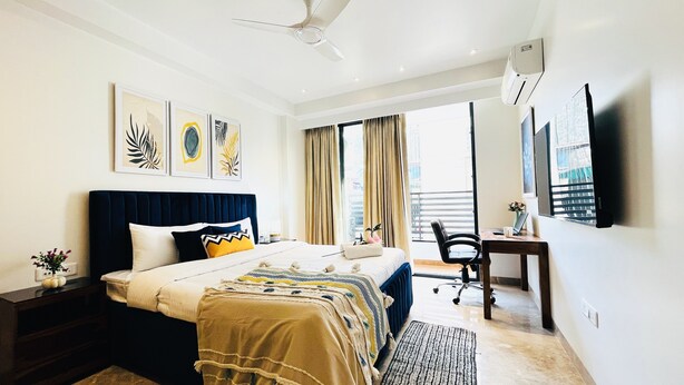 A hotel room with a bed, a desk, a chair, a ceiling fan, and artwork on the walls. - BluO 3BHK CityWalk Mall, MAX Saket -Lift, Balcony (New Delhi)