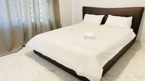 A neatly made bed with white linens and a folded towel, a dark headboard, and a window with sheer curtains.