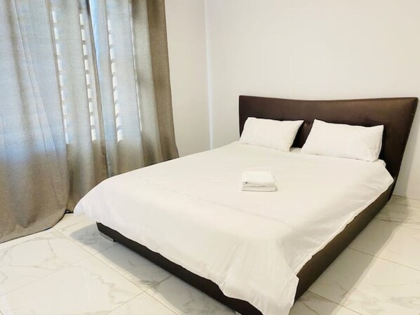 Una cama bien tendida con sábanas blancas y una toalla doblada, un cabecero oscuro y una ventana con cortinas traslúcidas.
