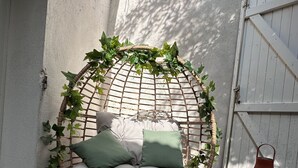A wicker swing chair with cushions, surrounded by greenery, in a shaded outdoor area with a white fence and a green folding chair.