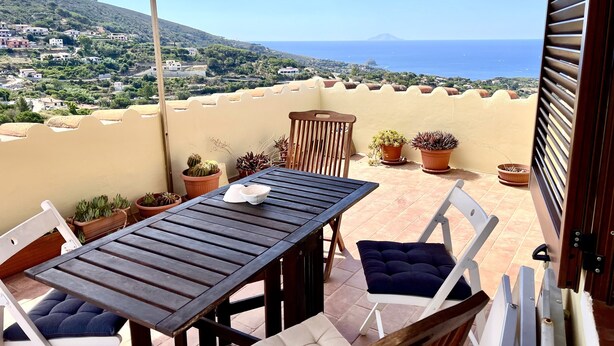 Una terraza con mesa y sillas, plantas en macetas y vistas al mar y colinas distantes.