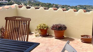 Una terraza con una silla de madera, dos plantas en macetas y vistas al mar y colinas lejanas.