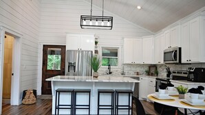 A modern kitchen with white cabinets, a large island with bar stools, stainless steel appliances, and a hanging light fixture. - Moonlight Rest-Sleeps up to 4/Hot Tub (Broken Bow)
