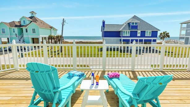 A wooden deck with turquoise Adirondack chairs and a small table, overlooking a beach with houses in the background. - Epic 6BR Beach View * Steps to Beach * Sleeps 20+ (Galveston)