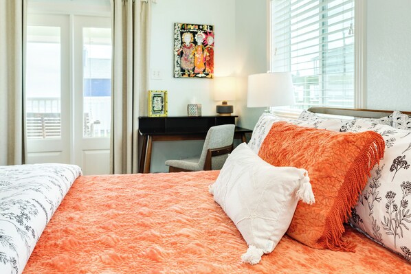 A hotel room with a bed, a desk, and a chair. There is a window with blinds, a picture on the wall, and a lamp on the desk.
