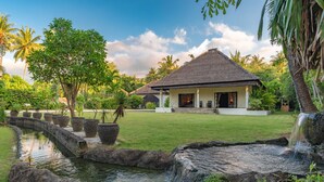 A house with a thatched roof, surrounded by a well-maintained garden with a small pond and a waterfall. - Villa Bukti Paradise (Kubutambahan)