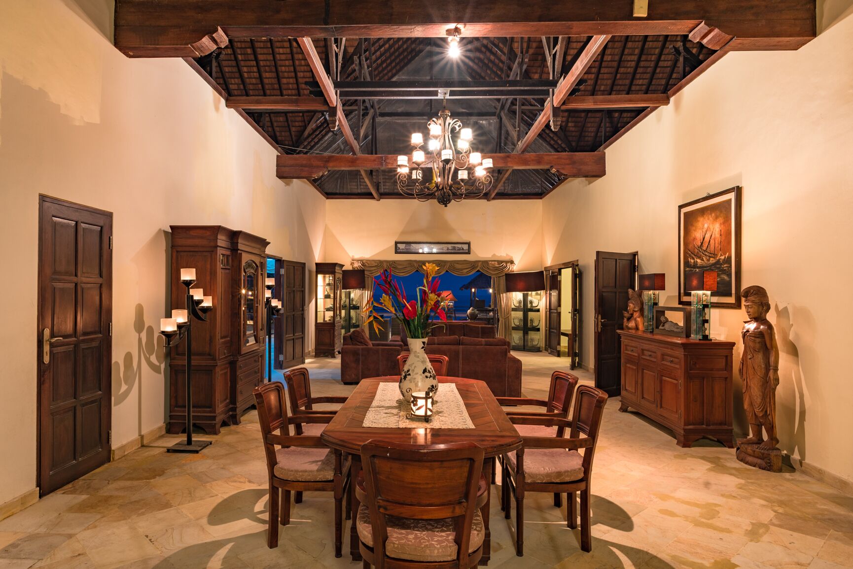 A dining area with a wooden table, chairs, and a chandelier. There is a statue and a painting on the wall.