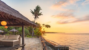 A beachfront resort with a thatched-roof lounge area, stone flooring, and a view of the sunset over the ocean. - Villa Bukti Paradise (Kubutambahan)
