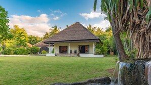 A house with a thatched roof, surrounded by a well-maintained lawn and a small waterfall. - Villa Bukti Paradise (Kubutambahan)