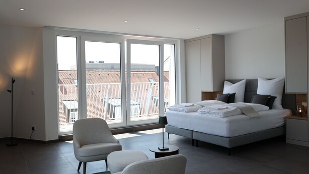 A modern hotel room with a large bed, a seating area, and a view of buildings through large windows. - Studio mit Küche 12 (München)