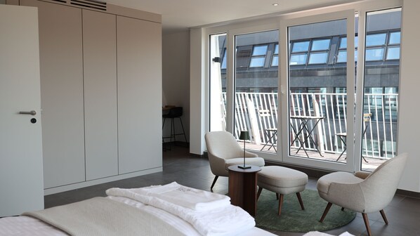 A modern hotel room with a large bed, two armchairs, a small table, and a balcony with a view of the city.